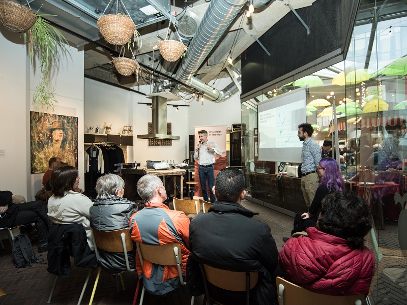 Aufnahme aus der Markthalle 8 in Bremen. Zwei Männer halten einen Vortrag zum Thema Marsbesiedelung. Im Vordergrund sitzen Zuhörer:innen auf Stühlen.