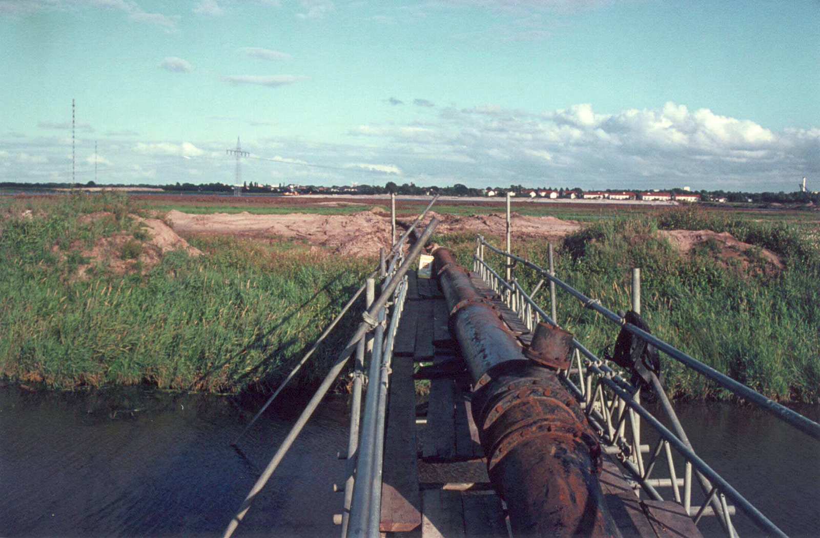 Im Vordergrund befindet sich eine rostige Rohrleitung, die über einen Graben verläuft. Ihr weiterer Verlauf verliert sich in der Wiese zwischen ein paar Sandhügeln. Am Horizont zeichnet sich die Häusersilhouette von Horn-Lehe ab.