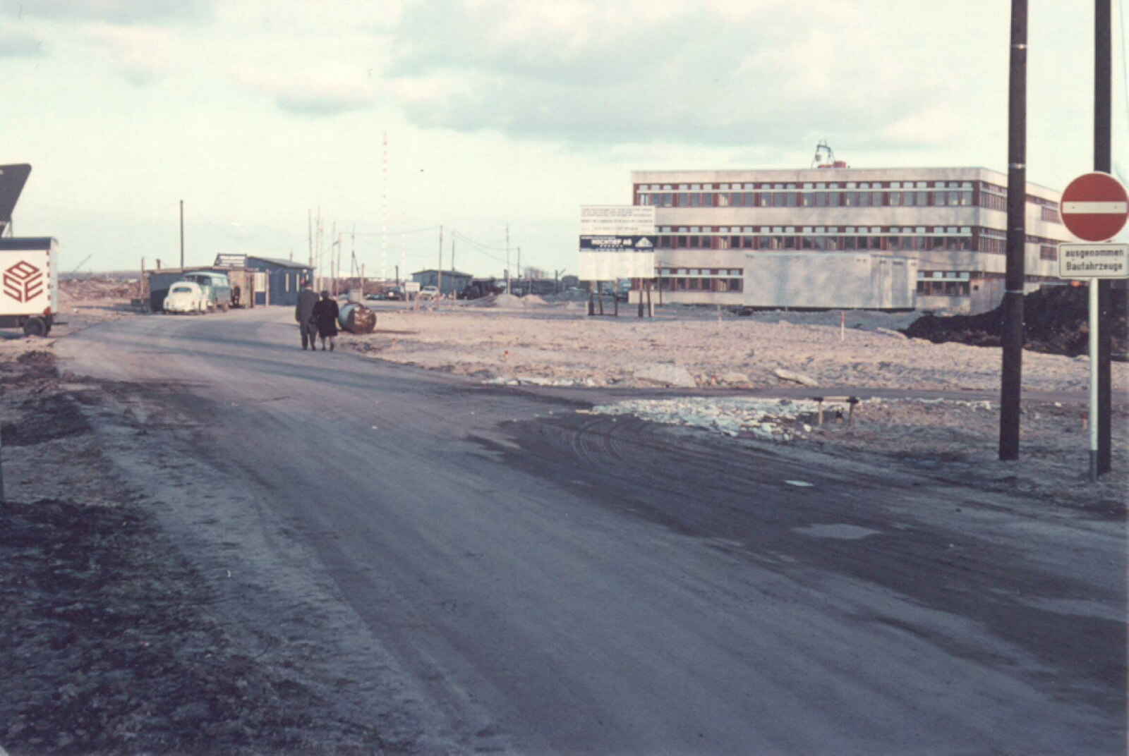 Blick auf das dreigeschossige Gebäude mit einer Bautafel davor. In der Verlängerung der Parkallee stehen Unterkünfte für Bauarbeiter. Die Straße ist nur zum Befahren für Baufahrzeuge zugelassen, davon kündet ein Schild am rechten Straßenrand. Ein Mann und