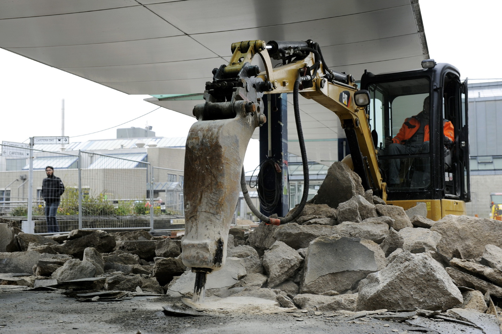 Ein Bagger reißt Betonboden am Boulevard auf, hinter dem Bauzaun geht ein Mann spazieren.