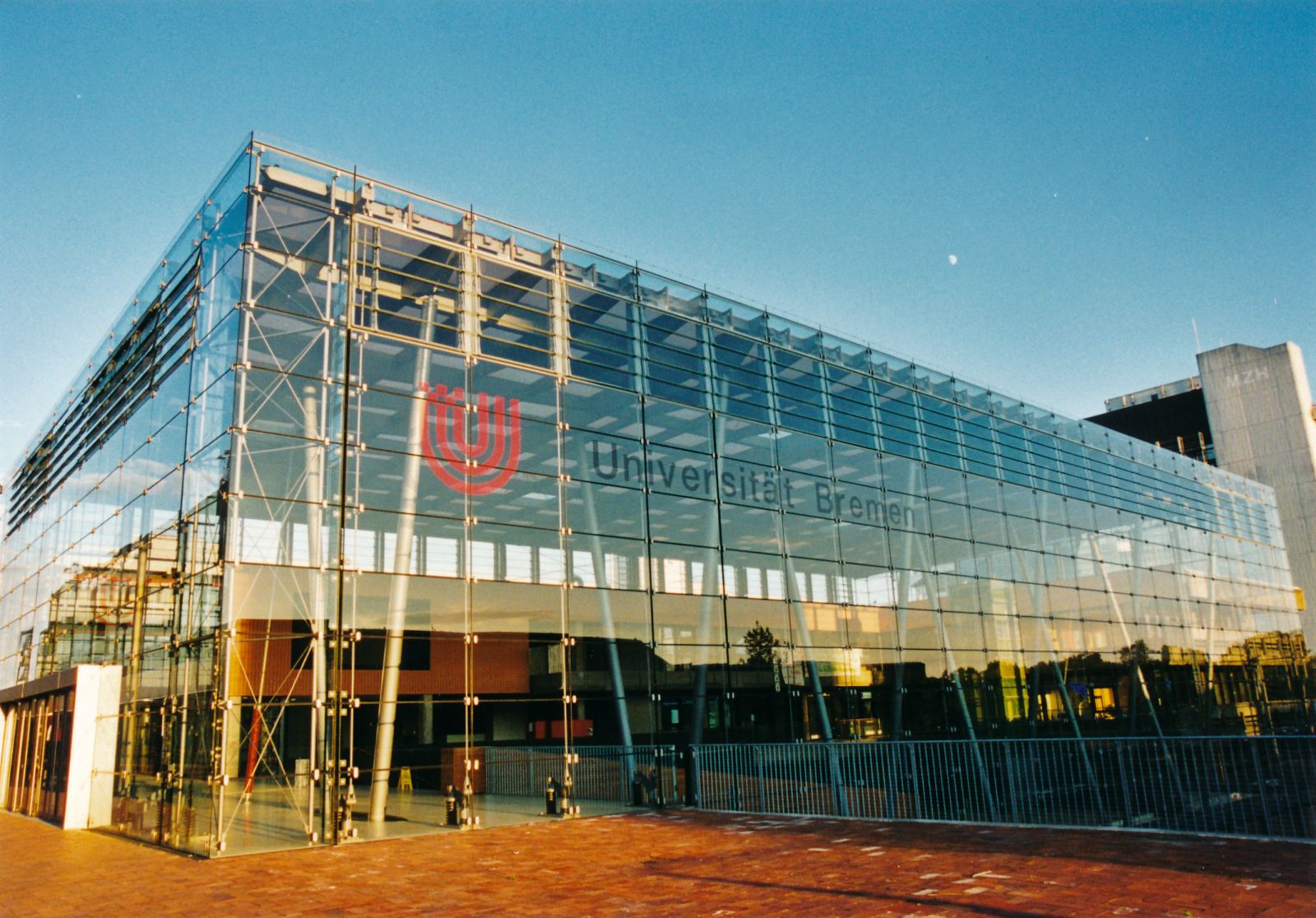 Auf dem Bild sieht man mittig ein großes rechteckiges Gebäude, bestehend aus vielen kleineren, rechteckigen Glaskacheln, das ist die Glashalle. Oben links an der Glasfront ist das Logo der Universität Bremen. Die Sonne scheint seitlich links auf das Gebäu