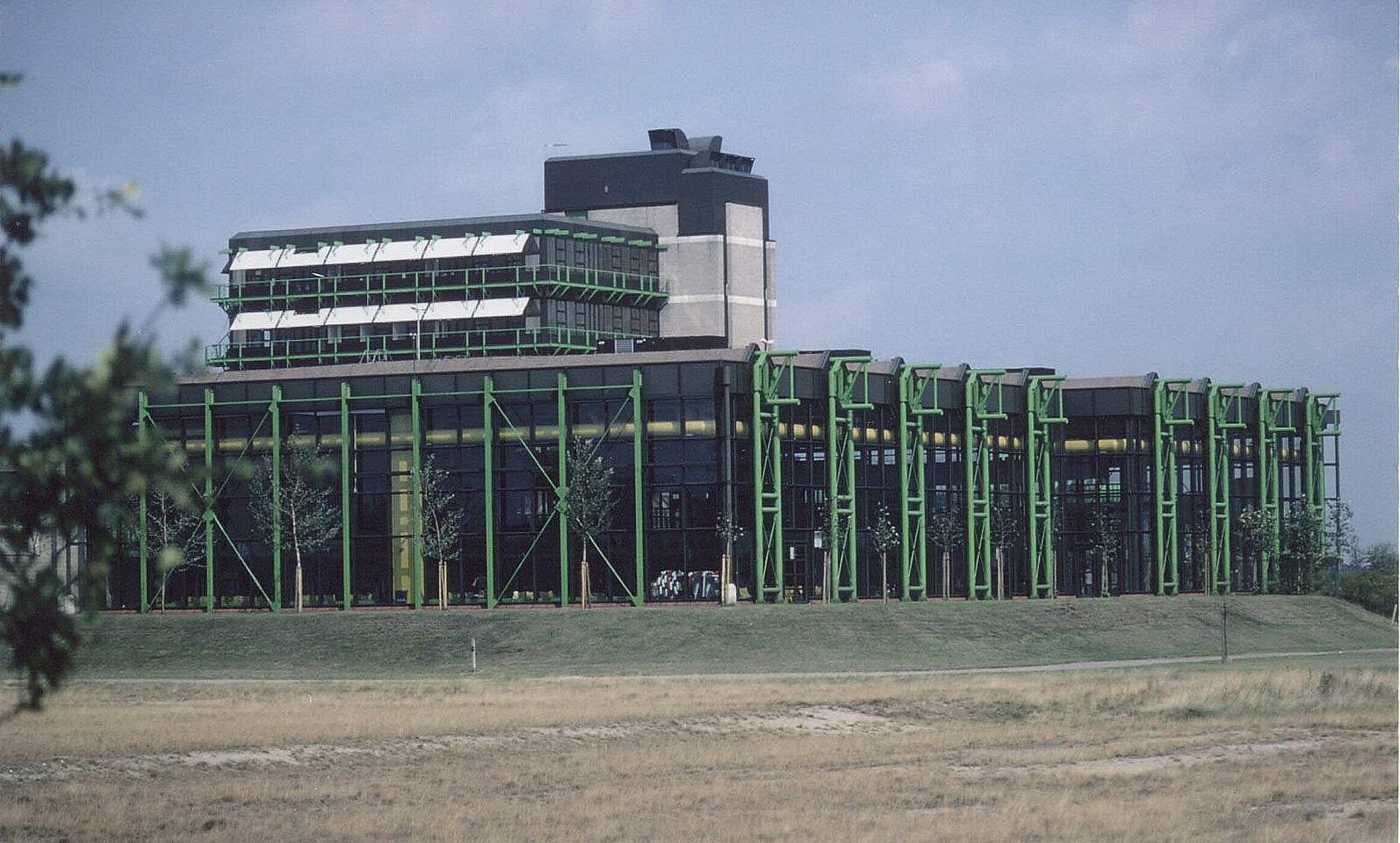 In der Bildmitte befindet sich ein umliegend verglastes, rechteckiges Gebäude mit einer grasgrünen außenliegenden Stützkonstruktion. Dieses futuristische Gebäude ist das Uni- Bad im Sportbereich der Universität Bremen. Im Bildvordergrund befindet sich ein