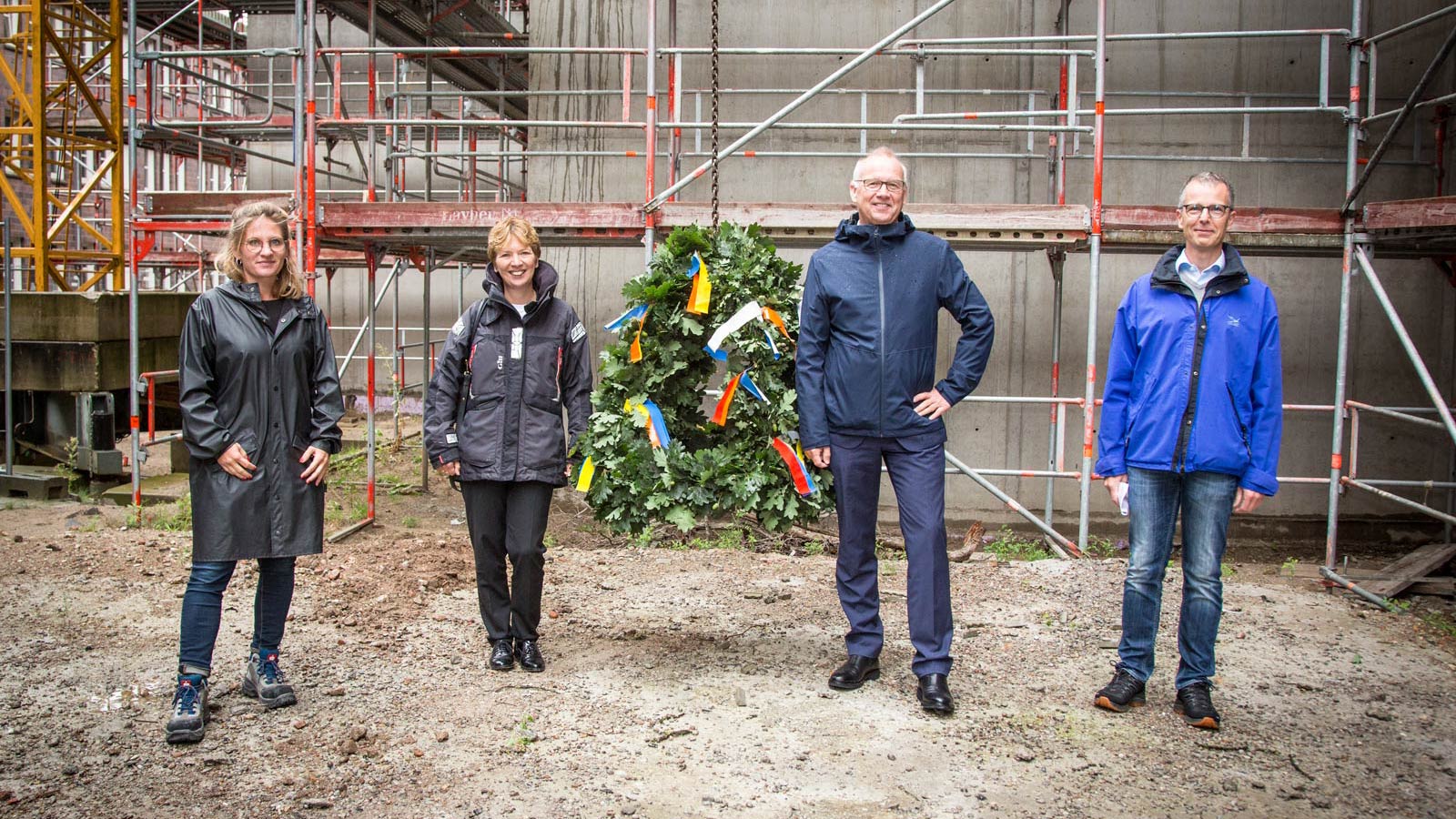 Vier Personen stehen auf einer Baustelle vor einem Richtkranz und schauen in die Kamera.