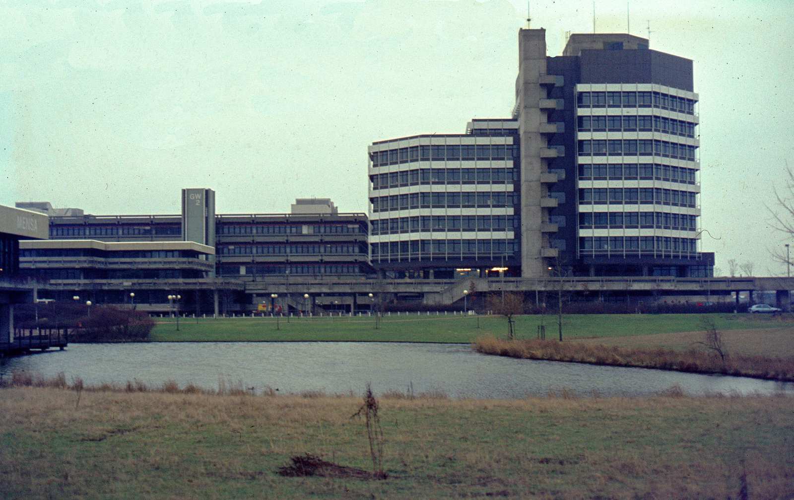 Blick von Westen auf das Mehrzweckhochhaus (MZH), links dahinter sind Teile des Gebäudes Geisteswissenschaften 2 und über die gesamte Breite des Bildes ein Teil des Boulevards abgebildet. Im Vordergrund ganz links ragt die Mensa ins Bild, in der Mitte des