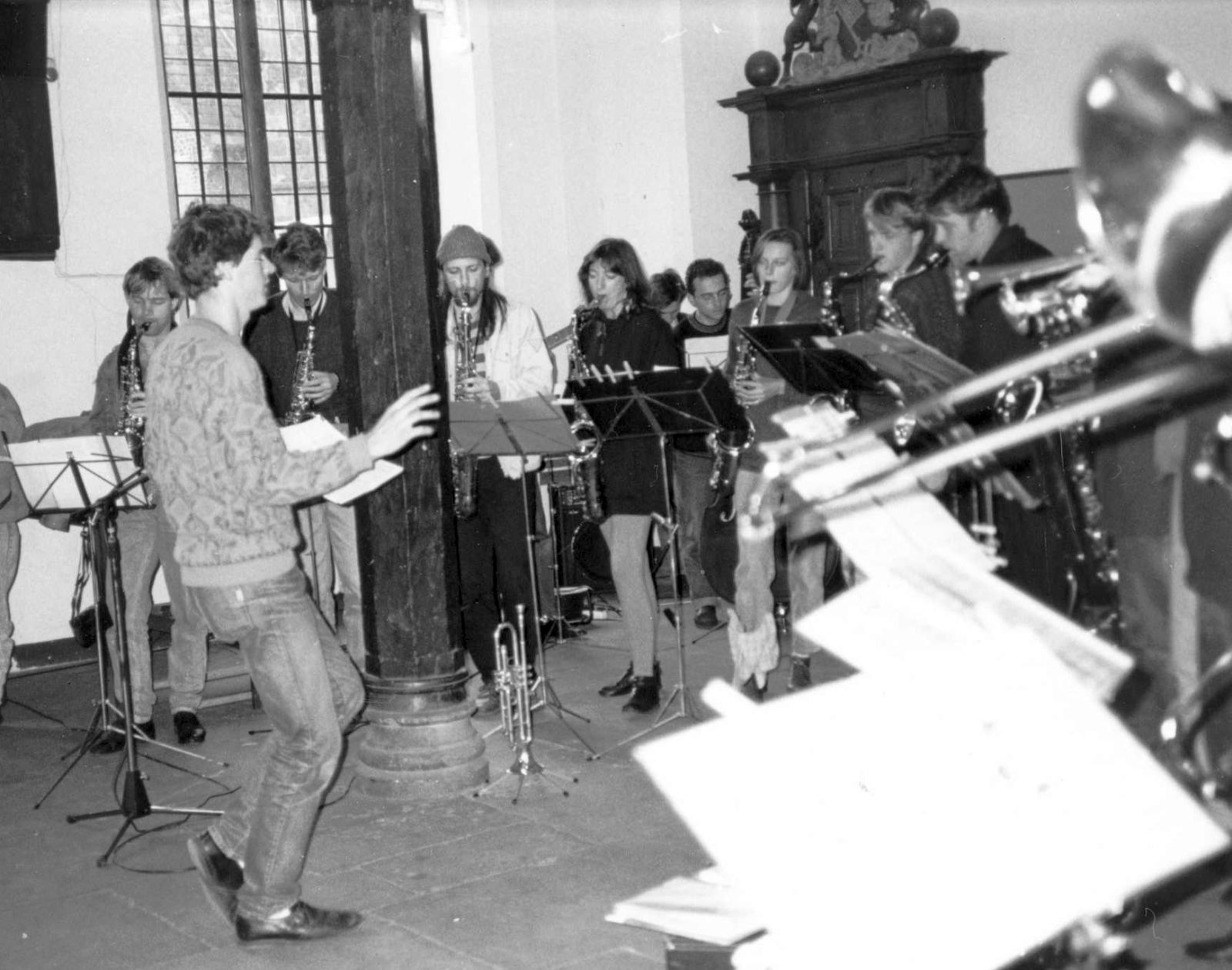 Schwarzweißaufnahme der Uni-Bigband während ihres Konzerts im Oktober 1991 in der Unteren Rathaushalle. Zu sehen sind in der Mitte der Dirigent und im Halbkreis um ihn herum die Blechbläser und -bläserinnen (vor allem Saxophon).
