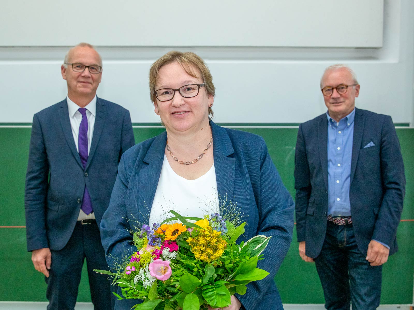 Frauke Mayer mit Blumenstrauß in den Händen. Hinter ihr stehen Rektor Bernd Scholz-Reiter und Altkanzler Martin Mehrtens.