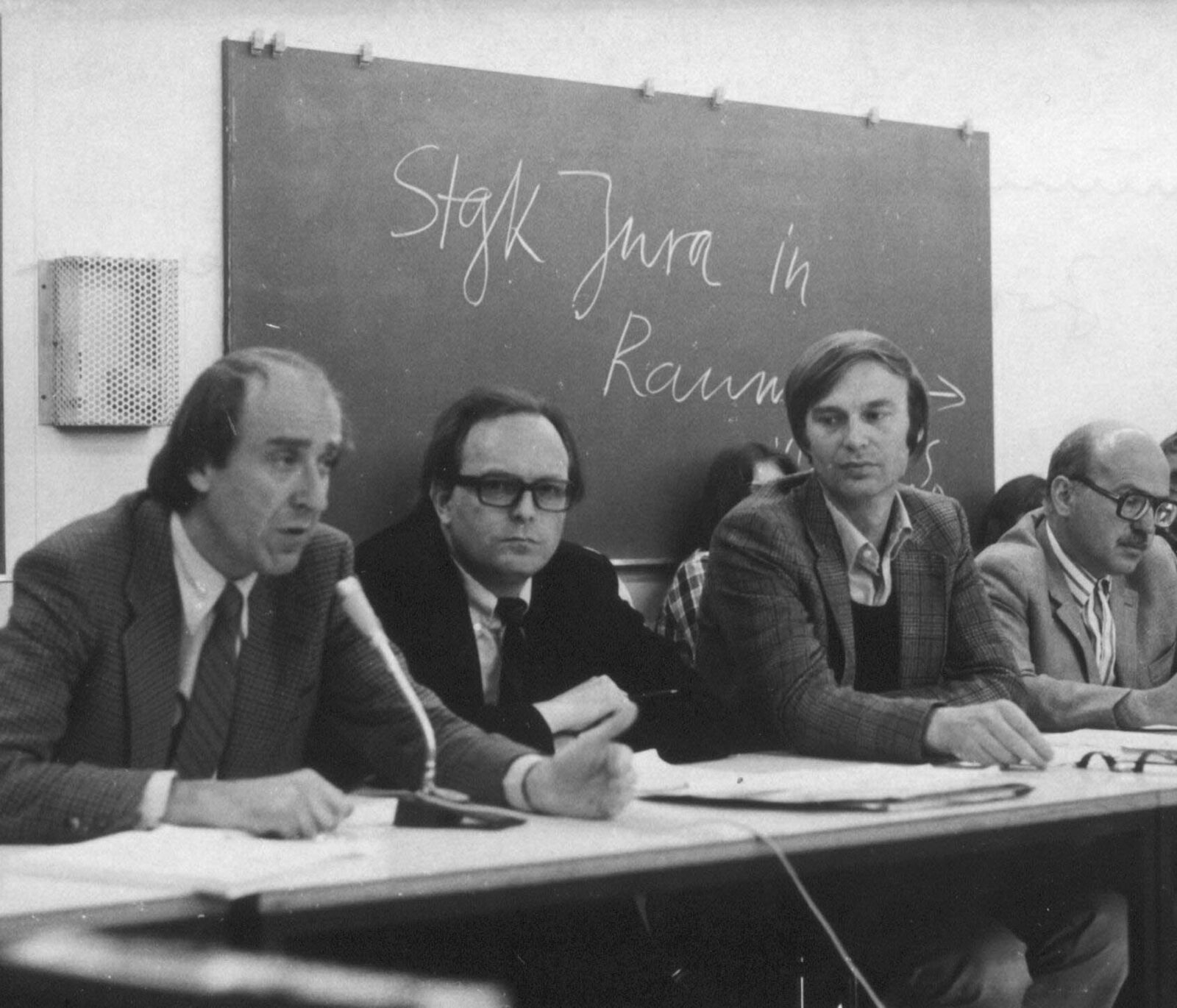 Gerhard Stuby sitzt mittig an einem Podium und hört konzentriert dem Redner zu (in diesem Bildausschnitt nicht zu sehen). Neben ihm sitzen der Uni-Rektor Thomas von der Vring (links) und der Bildungssenator Hans-Werner Franke (rechts). Herr Stuby trägt ei