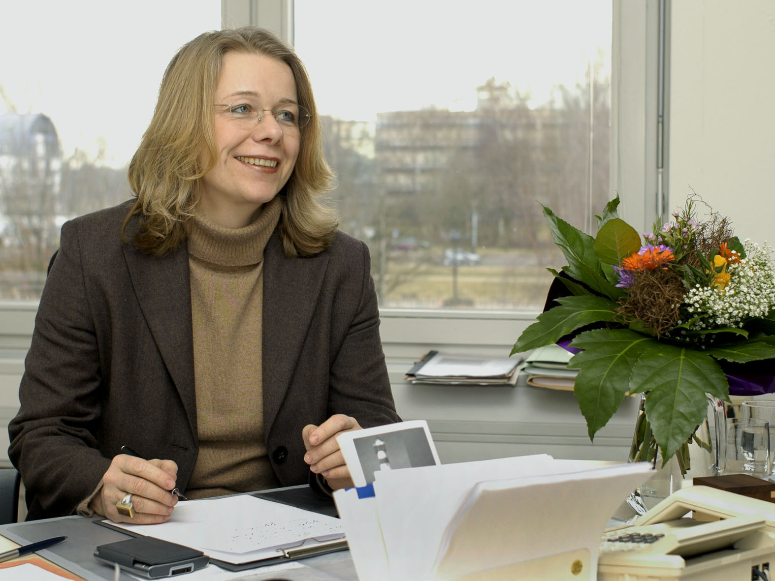 Eine Frau sitzt an einem Schreibtisch, hält einen Stift und lächelt.