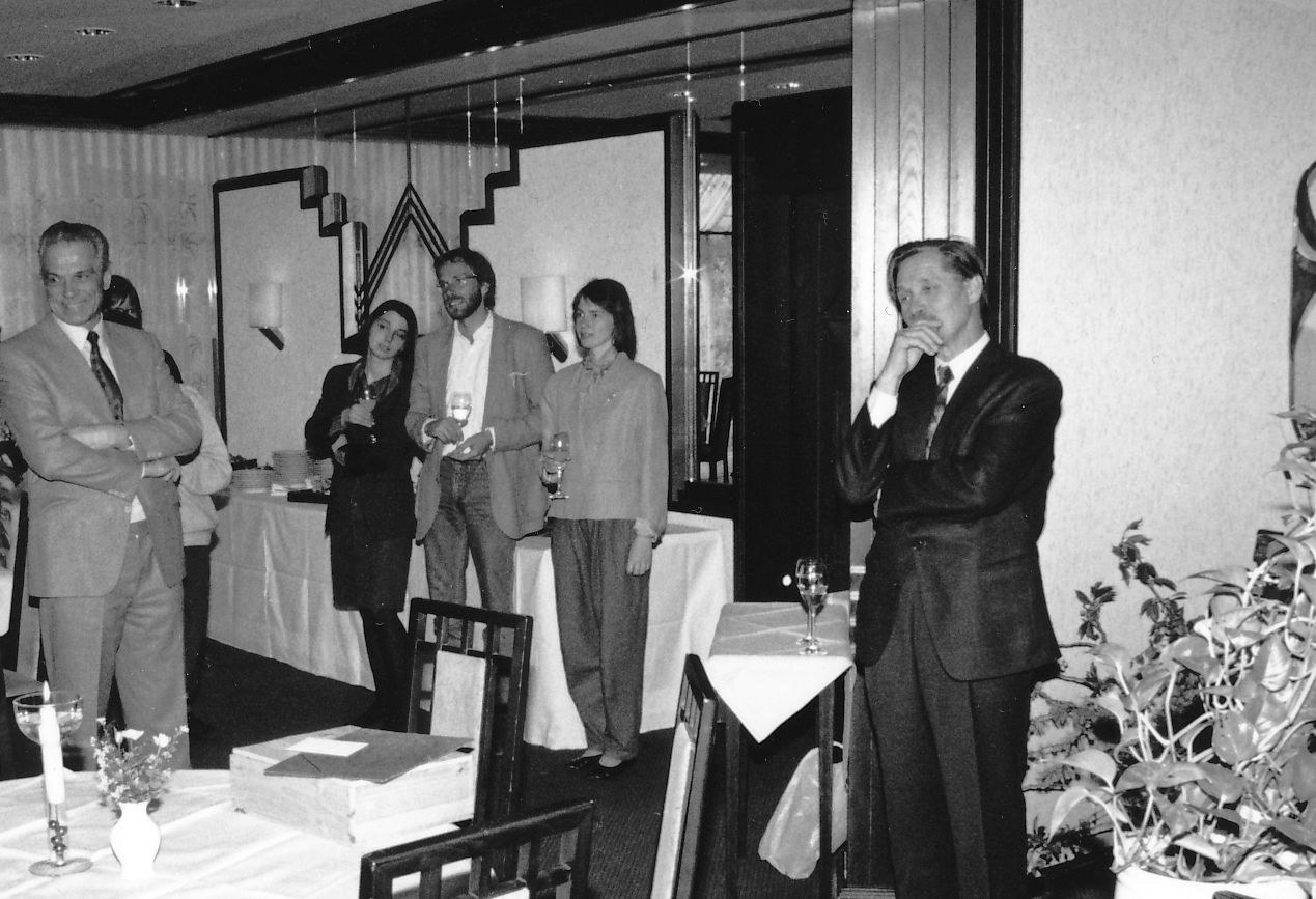 Das Foto ist während der Abschiedsfeier für Hermann Cordes entstanden. Er trägt einen dunklen Anzug, steht an die Wand gelehnt und sieht nachdenklich aus. Im Bildzentrum steht ein Tisch mit einer Kerze. Es sind mehrere Gäste zu sehen, die lächeln und Gläs