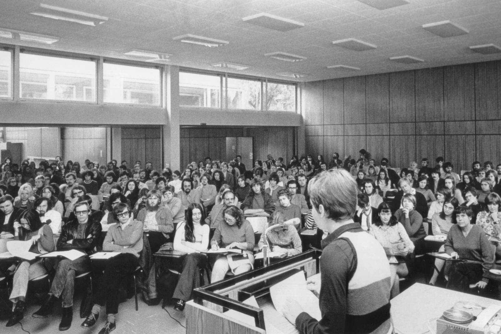 In einem großen Raum sitzen Studierende. Die Reihen sind dicht gefüllt. An einem Pult steht eine Frau.